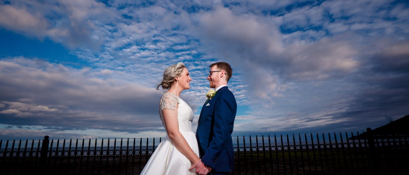 Bride and groom holding hands, smiling at each other against a dramatic sky with clouds, showcasing natural-light wedding photography in Northern Ireland.