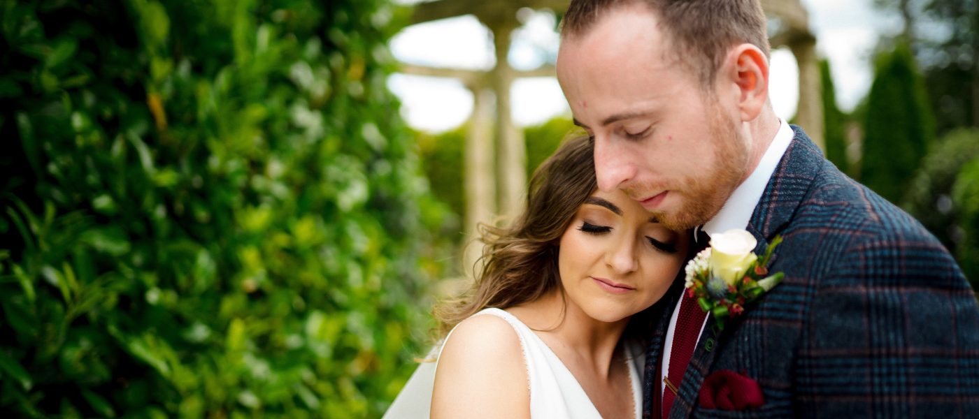 Couple embracing in a romantic pose amidst lush greenery at Tullyglass House Hotel, capturing the elegance and natural beauty of the wedding venue.