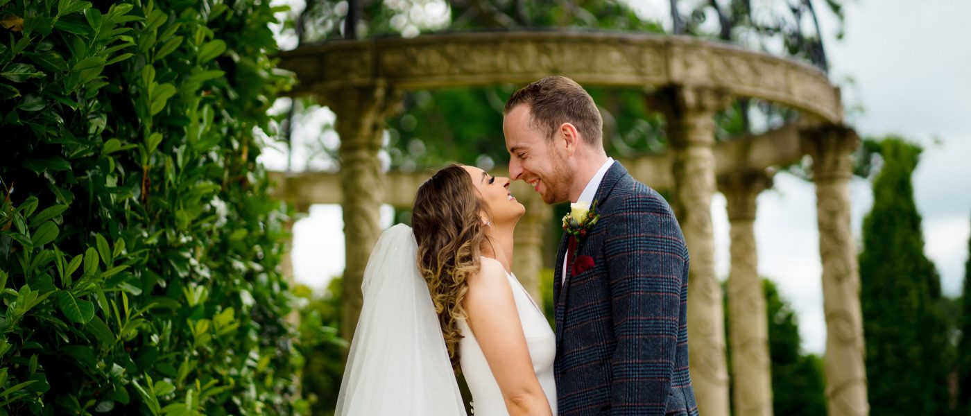 Bride and groom sharing a joyful moment under a classic stone gazebo surrounded by lush greenery at Tullyglass House Hotel, ideal for wedding photography.